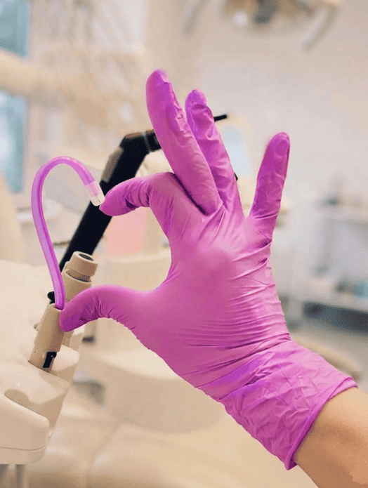 Vista en primer plano de la mano de un profesional de la salud, enfundada en un guante de nitrilo de color rosa brillante. La mano sostiene y dirige un tubo de succión dental de color morado translúcido, que está curvado en forma de corazón hacia el conector del equipo dental. La mano hace el gesto de formar corazón con el tubo con los dedos índice y pulgar alrededor de la base del tubo. El fondo es una consulta dental limpia y desenfocada.